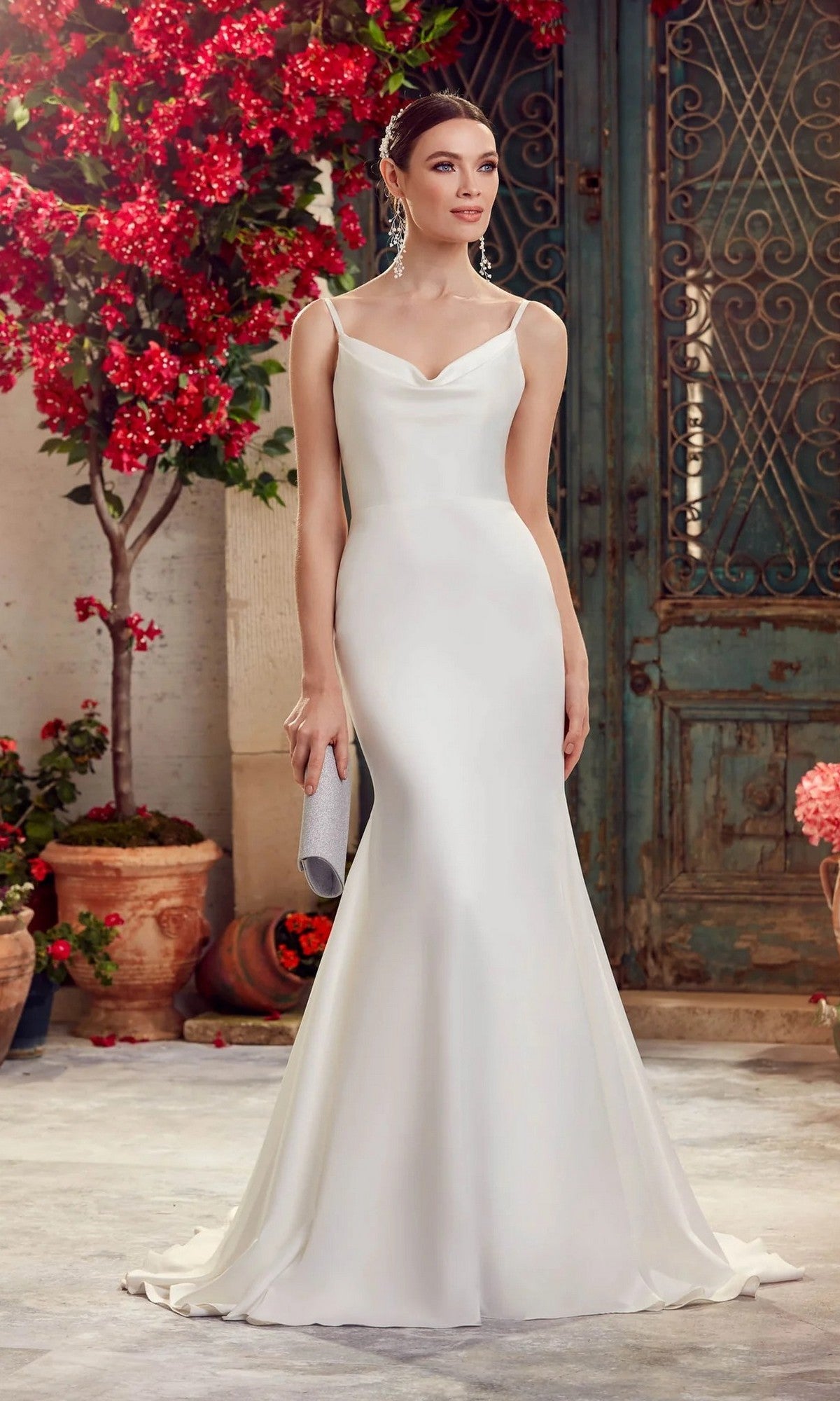 A woman in the Alyce 7051 Long White Dress, featuring a sleek fitted silhouette and cowl neckline in diamond white, stands outdoors holding a clutch. Red flowers and a rustic green door complete the scene.